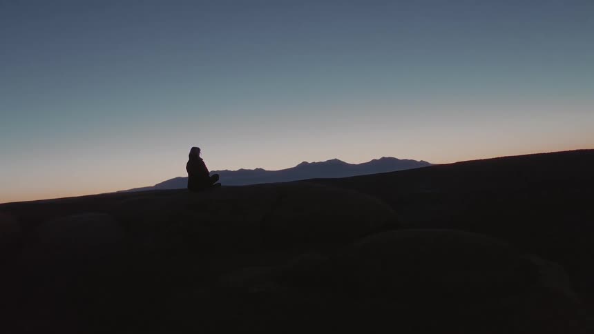 A woman sits with her legs crossed on a cliff in the mountains at sunset