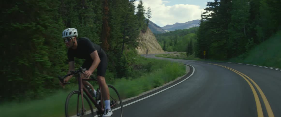 A cyclist rides a bicycle along a road through a countryside covered in mountain and forest during the day