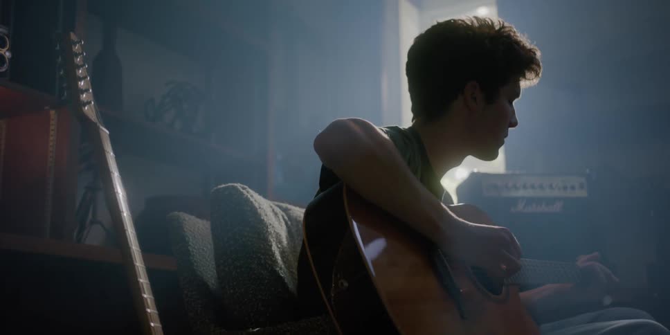 A man is sitting in a room playing an acoustic guitar and there is an electric guitar next to him as light shines brightly through the window
