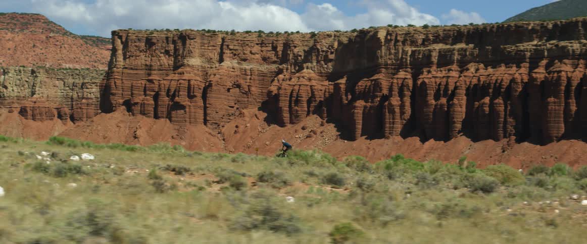 Vehicles pass a cyclist riding on a highway as a mesa sits in the distance on a sunny day