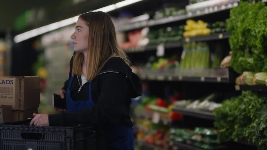 A worker in a grocery stores leans and looks bored as she pulls out her cellphone to send a text