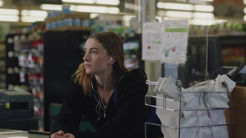 A cashier is leaning on the belt at a register in a grocery store as she listens on her earbuds