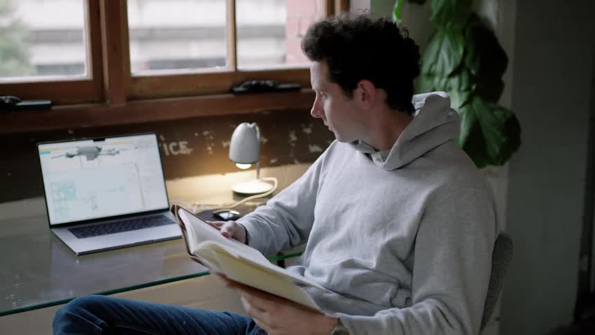 A man is reading a notebook and using his laptop