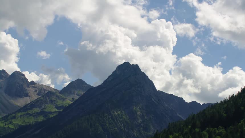 Clouds loom in the bright sky over a mountain range covered in forest