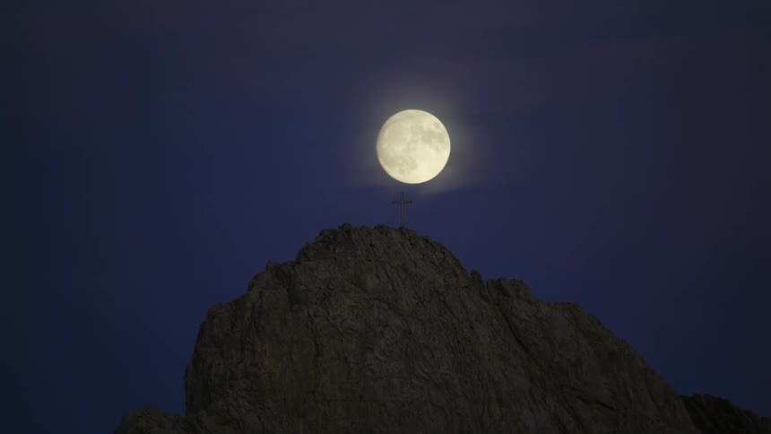 The full moon rises over a mountain and shines brightly behind a cross on the mountaintop as it moves higher in the night sky