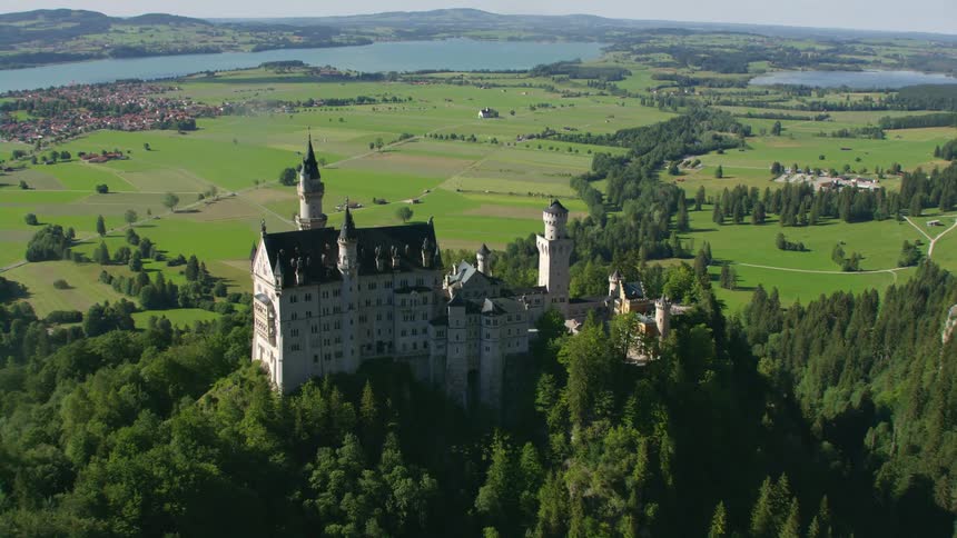 A castle atop the peak of a forested mountain overlooking a flat countryside