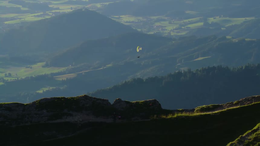 A person paragliding sails past a jagged mountain ridge covered in grass