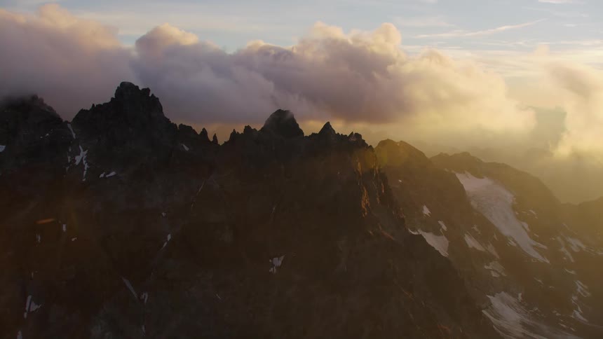 Thick clouds loom over a rocky mountain ridge covered in patches of snow during the day