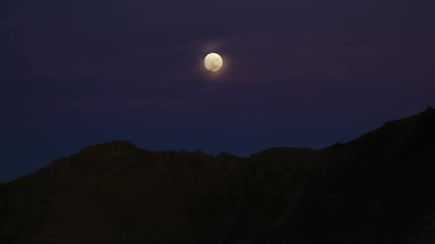The full moon looms in the cloudy sky over a mountainous countryside at night