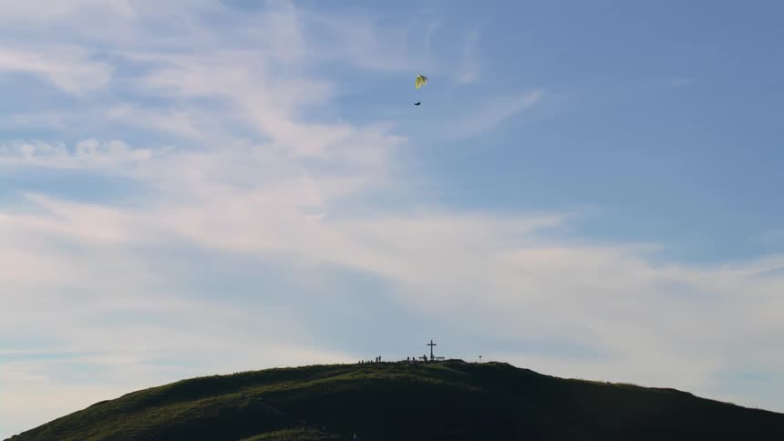 A person is paragliding over a mountain with a cross and a small cemetery on top of it
