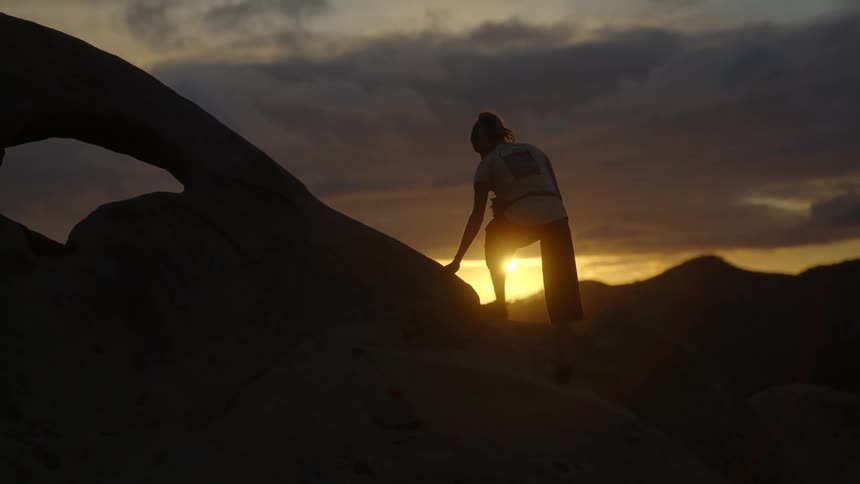 A woman climbs a mountain and stands at the top to look at the sun setting above mountains in the distance