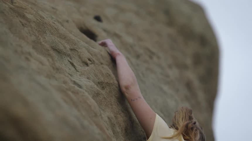 A woman rock climbing reaches her hand up for a hold and looks down