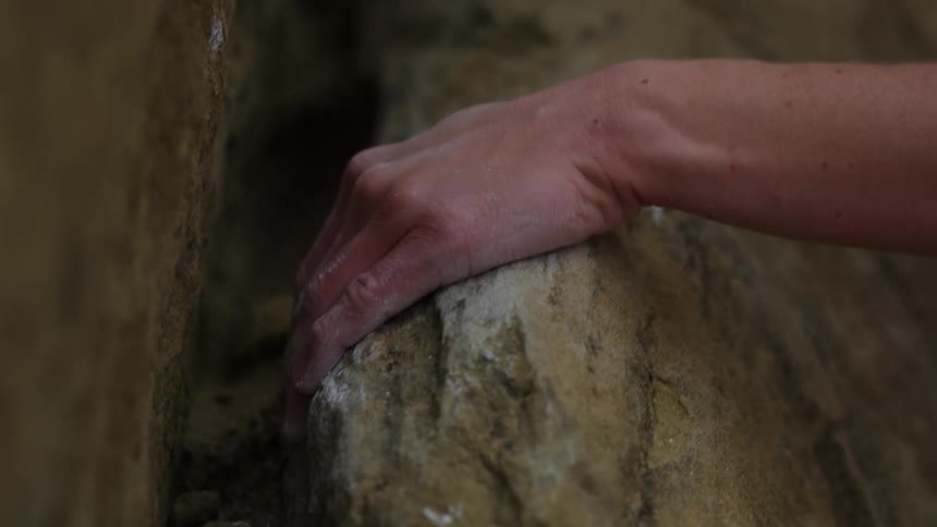 A person climbing a rock face grips a rock surface with a hand covered in chalk