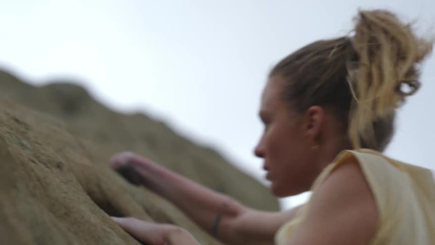 A woman is climbing up the side of a mountain and as she reaches up to grab ahold of the ledge above she looks down