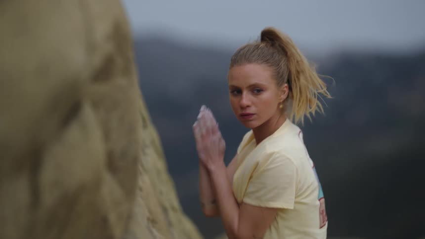 A blonde female rock climber stands in front of a rock and retrieves the chalk powder and rubs it all over her hands