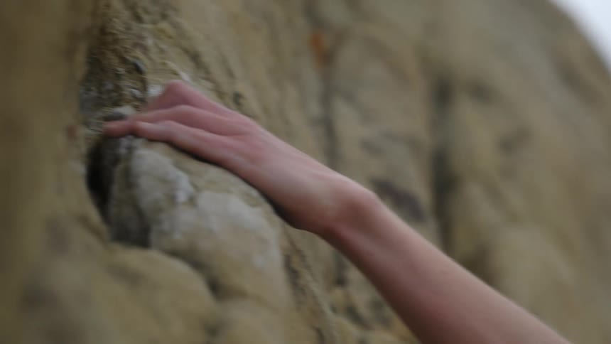 Blonde woman with a pony tail grabs onto a rock with her hand as climbs up the rocks on a mountain