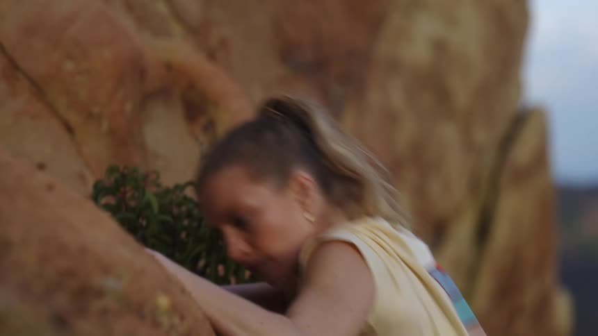Blonde woman with a pony tail and wearing a belt with a pouch climbs up the rocks on the side of a mountain
