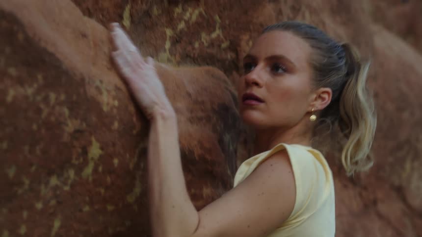 Blonde woman with an earring and a pony tail reaches with her arm as she climbs up a rock on the side of a mountain
