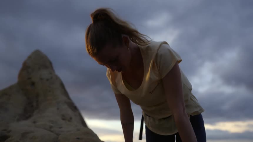 A woman climbs to the top of a natural rock face under a cloudy daytime sky