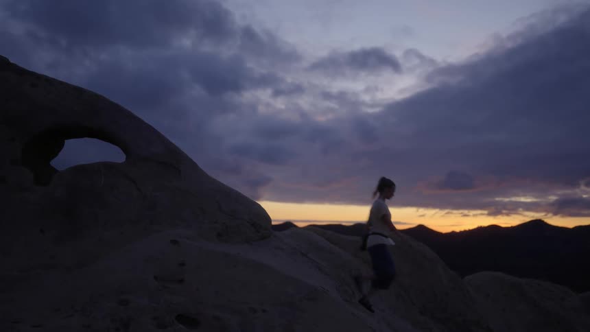 A woman walks down a slope of rock atop a mountain under a cloudy sky at sunset