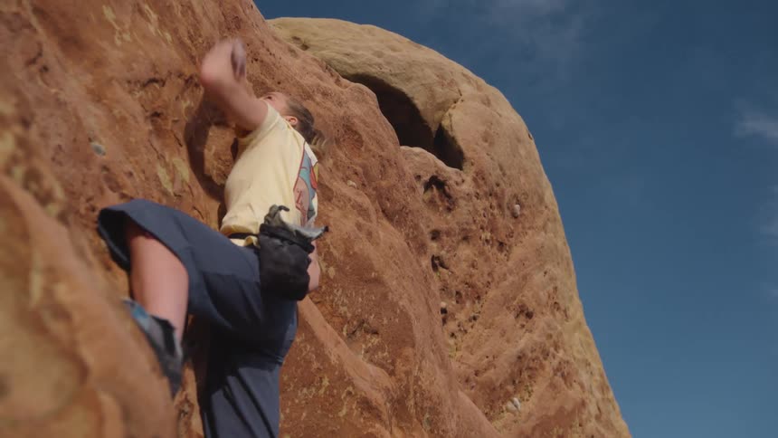 Blonde woman reaches with her arm while climbing up the rocks on the side of a mountain under a blue sky