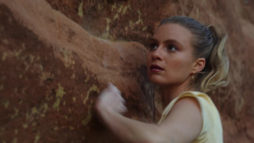 Blonde woman with a pony tail looks down then reaches with her arm to climb a rock