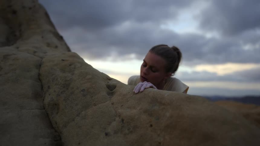 A woman is climbing the side of a mountain and her hand reaches up to the edge of a cliff and she pulls herself up