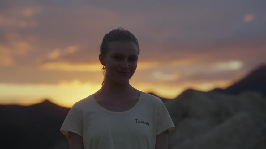 A woman standing atop a mountain raises her head and smiles while looking into the camera at sunset