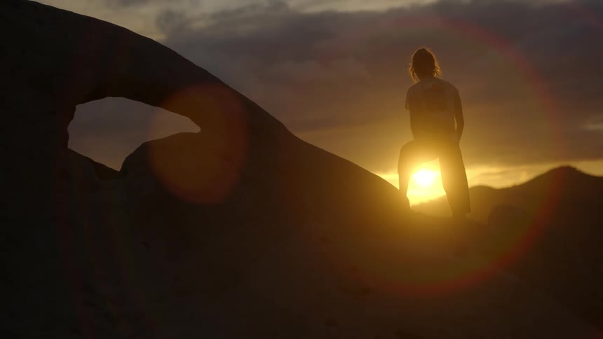 Woman with a pony tail climbs up to the top of the rocks and stops and poses as the sun rises