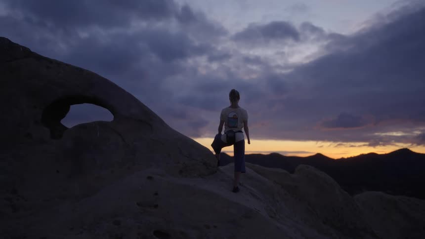 A woman stands on a mountain and looks at the sunset sky above mountains in the distance