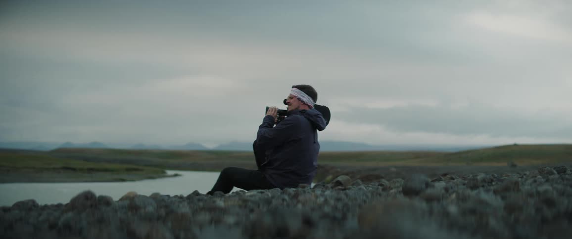 A man wearing a white headband is sitting along the side of a stream with a movie camera on his shoulder with mountains in the distance