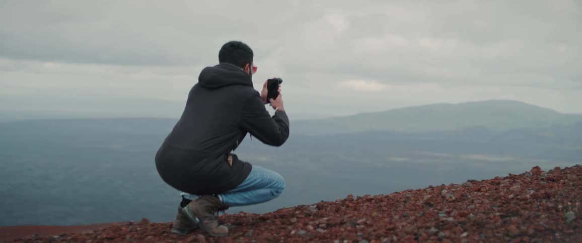 A man kneels down at the edge of a cliff to take a photo of nearby mountains