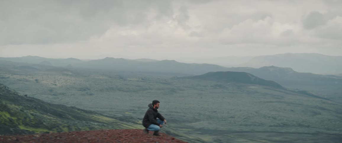 Young man wearing sunglasses crouching atop a mountain looking at a valley