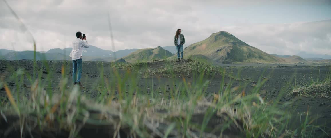 A woman is standing on a small mound in a field with mountains behind her as a man takes her picture