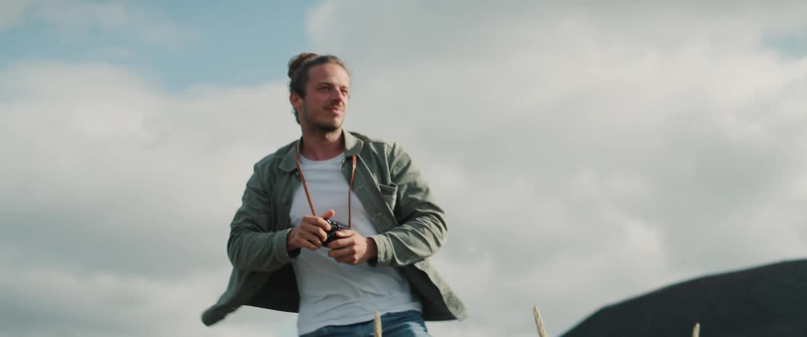 Male photographer with a bun green jacket white top and jeans stands up looking around in the field with mountain in the background
