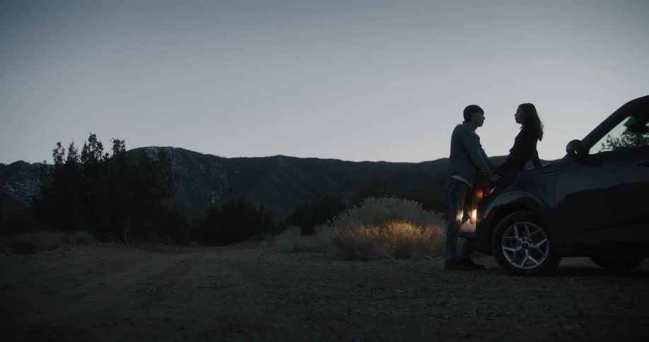 A woman sits on the hood of a car and laughs with a man who rests his hands on her legs