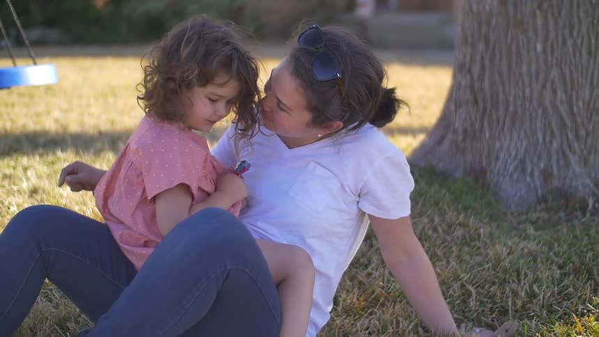 A girl is sitting on a woman's lap as she talks to her on the grass