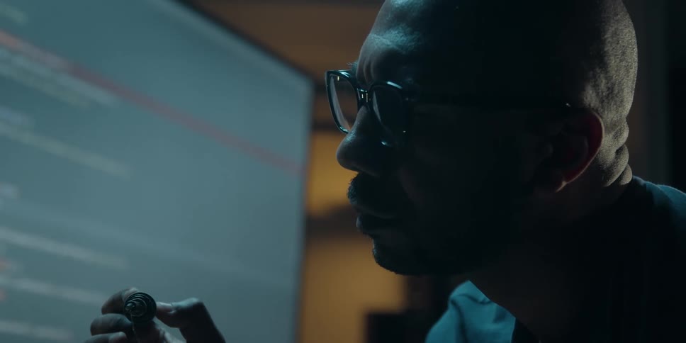 A man sits in front of a computer in a dark office and inspects a small tool