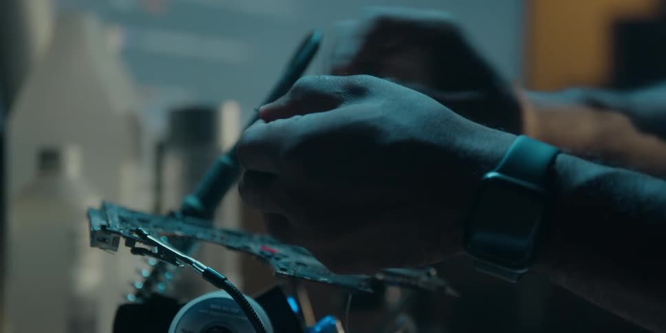A man is sitting at a table working on an electronic component and he cuts a piece of wire and twists the ends