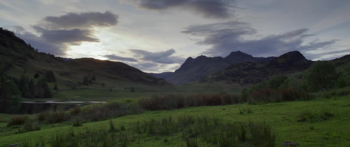 Small pond sits at the base of a mountain near the green field under a cloudy sky