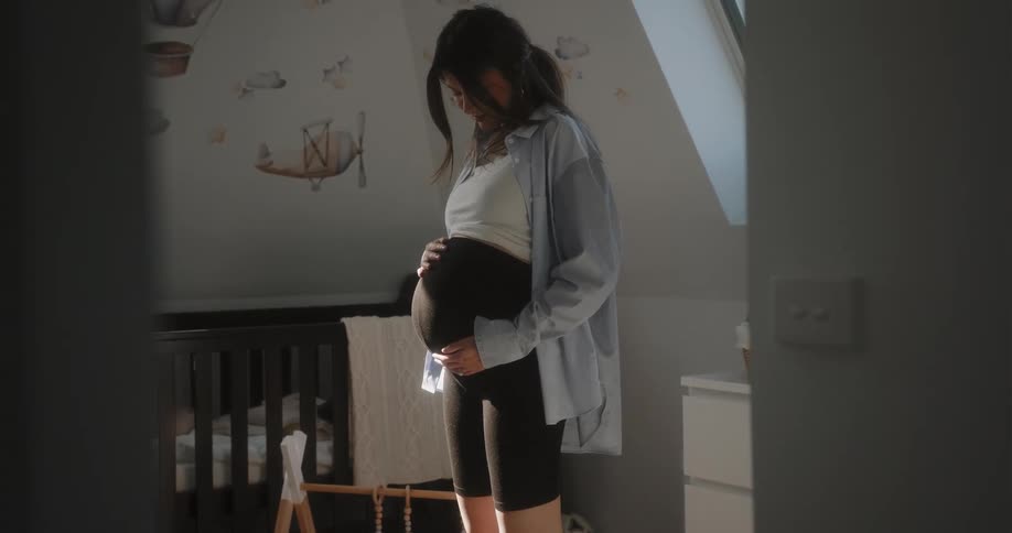 Pregnant woman with black shorts blue and white shirts touches her belly in baby room