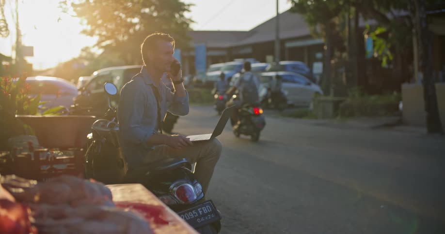 Man with blue shirt and brown pants sits on moped using laptop and talking on smartphone