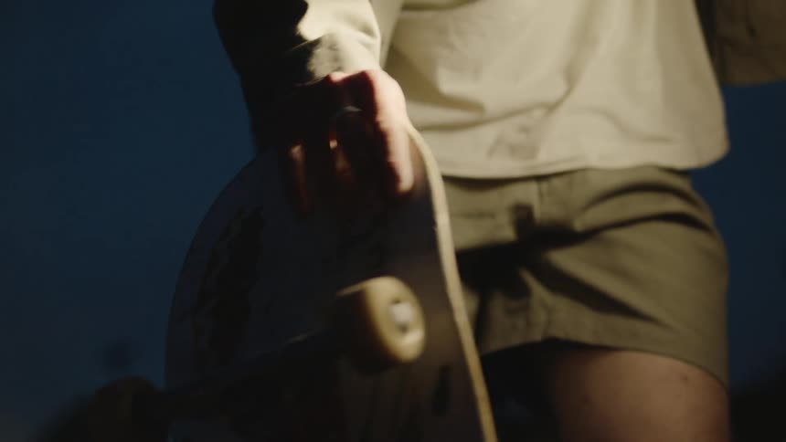 A teenage boy is standing in outside holding his skateboard and turning his wrist