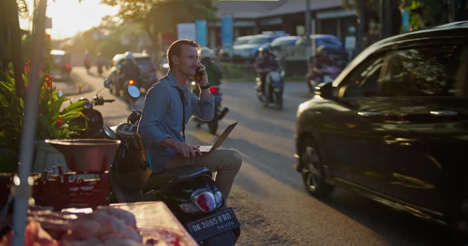 Man sitting on a parked motorcycle a the side of a busy street working on a laptop at evening