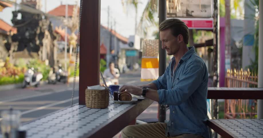 A man sitting at the counter of an outdoor tropical cafe typing on a laptop as cars and moped travel past