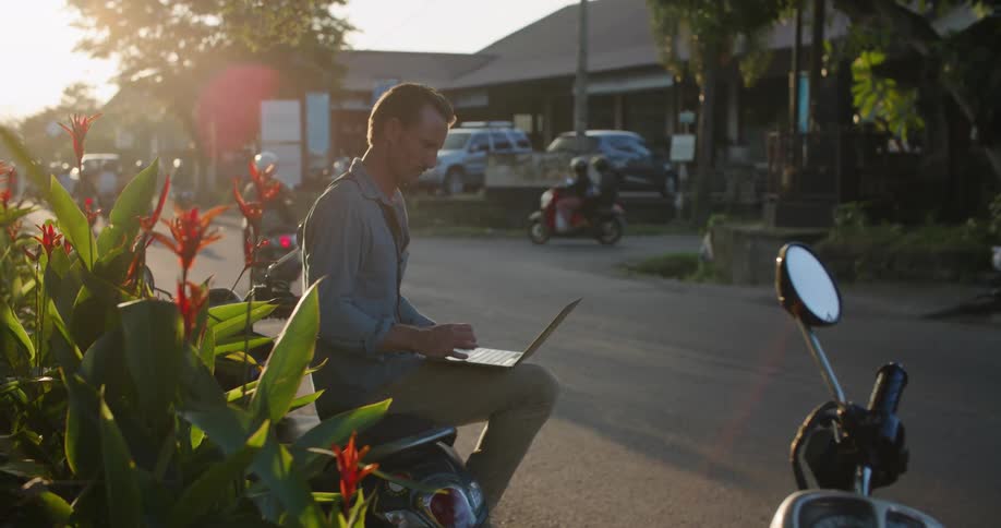 Man sitting on a parked motorcycle a the side of a busy street working on a laptop at evening