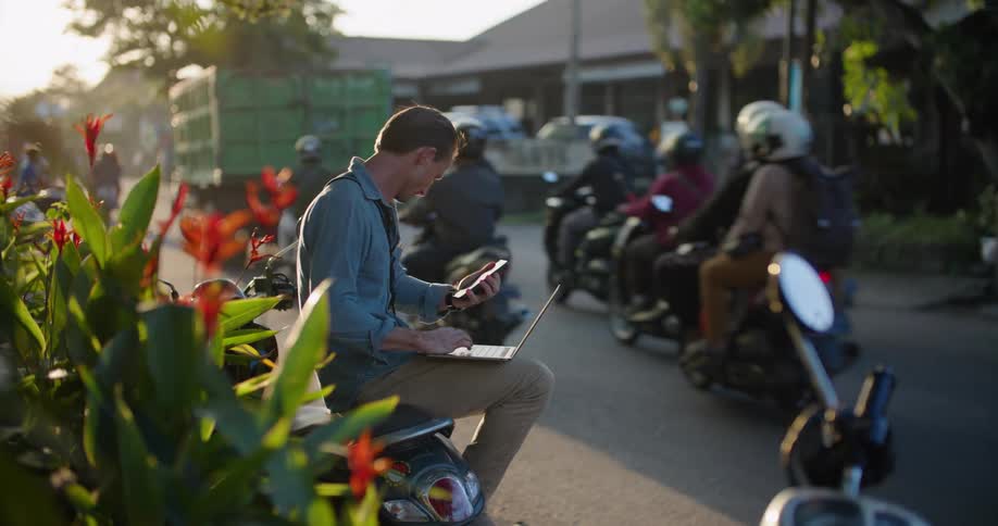 Man sitting on a parked motorcycle a the side of a busy street working on a laptop at evening