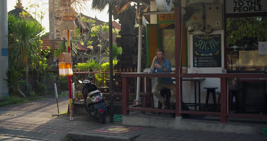 An older man is sitting outside by a store talking on the phone