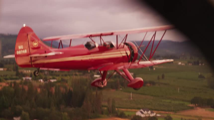 Aerial view of a red biplane flying over a small town near a mountain at morning