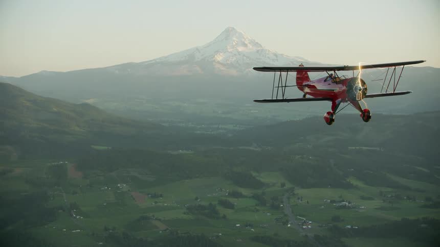 The propeller spins on a red airplane as it flies over the valley near the snow covered mountain